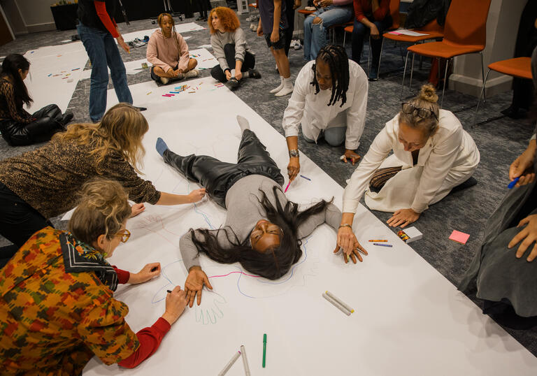 A person lies on the floor on a large piece of white paper. A group of people draw around them using a range of pens and pencils. 