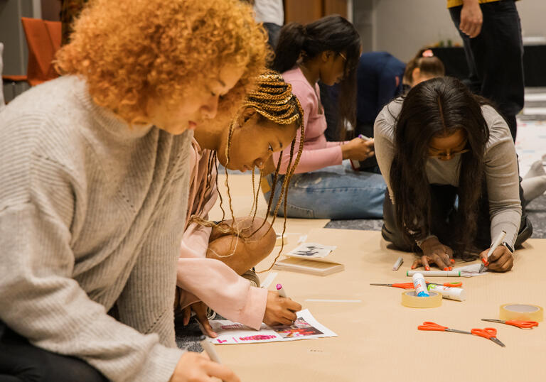 A group of people sit beside each other on the floor, drawing, colouring and collaging.