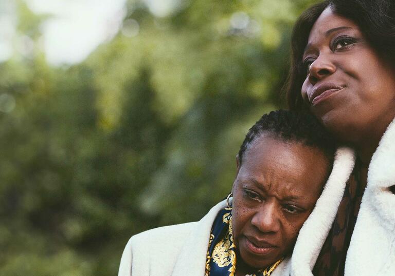 Two women in a park - one resting her head on the shoulder of the other.