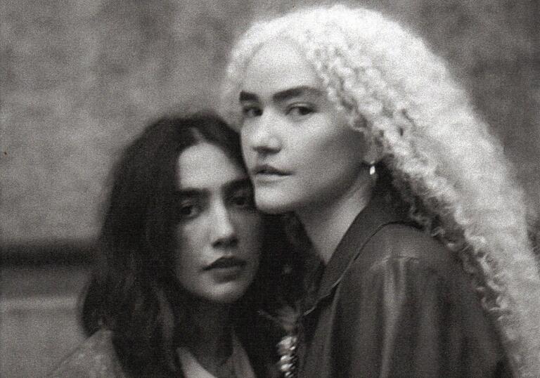 The musical duo Anavitória with Ana Clara Caetano Costa and Vitória Fernandes Falcão posing as they face each other.