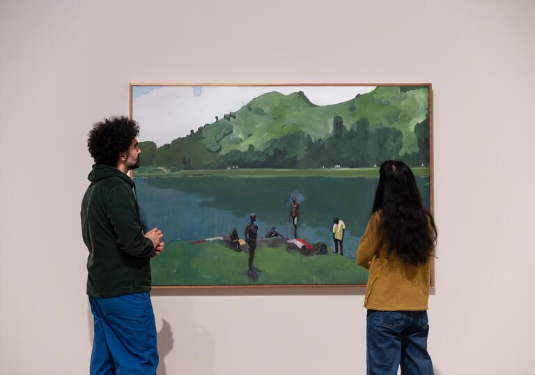A man and a woman stand in front of a large painting depicting people lounging on the grass next to a large body of water.