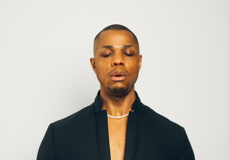 Davone Tines wearing a black suit with his eyes closed and hands in front of his chest