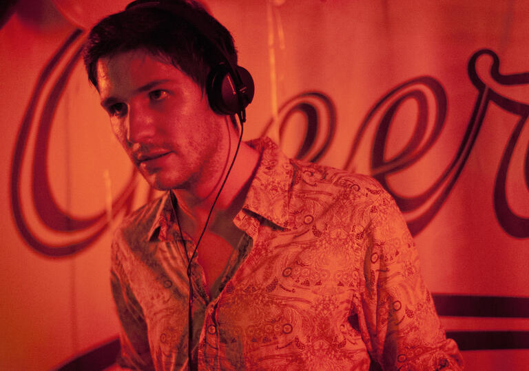 A white, French man with brown hair stands in a DJ booth, covered in red lights