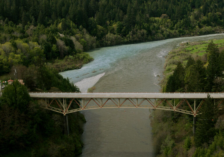 A still from Lucy Raven's moving image installation called Murderers Bar. It shows a bridge over a large river, surrounded by tree-covered hills.