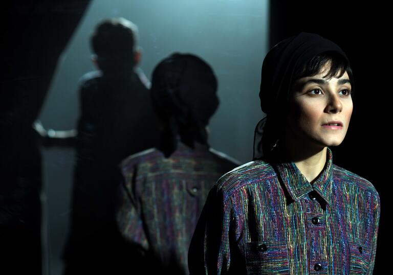 A woman is lit in the foreground is reflected by a mirror, as someone else in shadow walks through a curtain. 