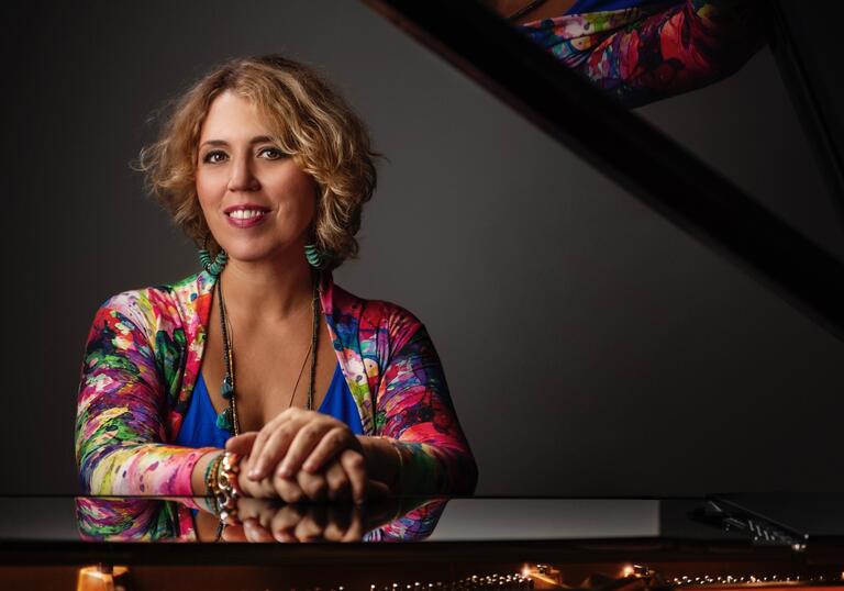 Gabriela Montero wearing a colourful dress sitting by a piano