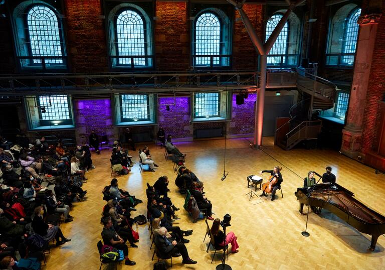 A view of the Jerwood Hall in LSO St Luke's from the balcony, looking down on rows of audience members sitting on chairs, facing a cellist and pianist on stage