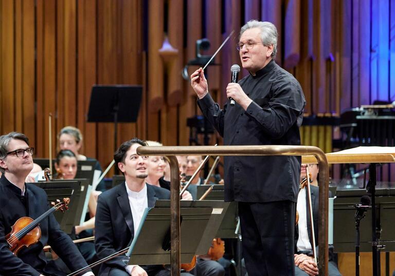 Sir Antonio Pappano standing on the podium facing the audience, talking to the audience, with the London Symphony Orchestra sat on-stage behind him