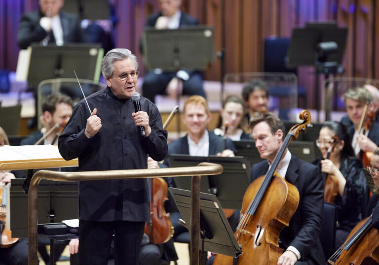 Sir Antonio Pappano standing on the podium on the Barbican stage, facing the audience and talking into a microphone
