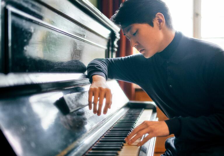 Seong-Jin Cho sat at a piano, facing towards the keys, with his arm resting on the lid of the instrument