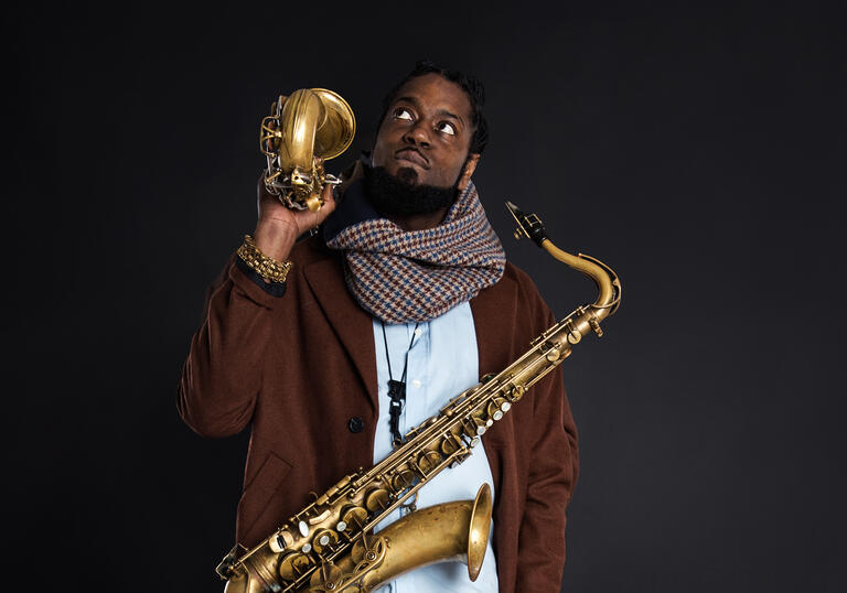 Soweto Kinch against a dark background, holding two saxophones