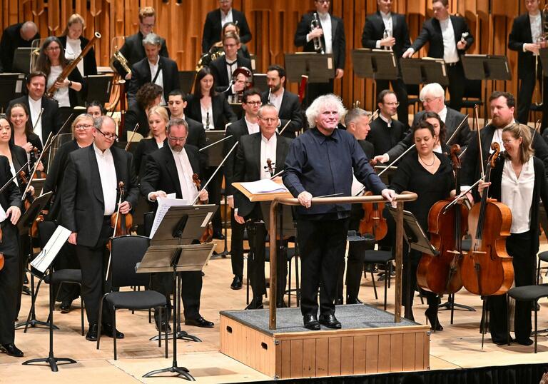 Sir Simon Rattle standing on the podium on the Barbican stage, facing the audience and smiling, with the London Symphony Orchestra standing behind him