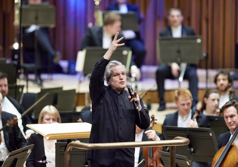 Sir Antonio Pappano standing on the conductor's podium, facing the audience, talking into a microphone with his right-hand raised