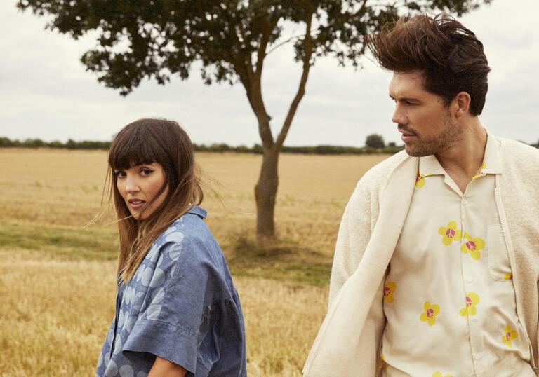 An image of both members of Oh Wonder walking away from each other in a field of grass with a tree behind them in the distance