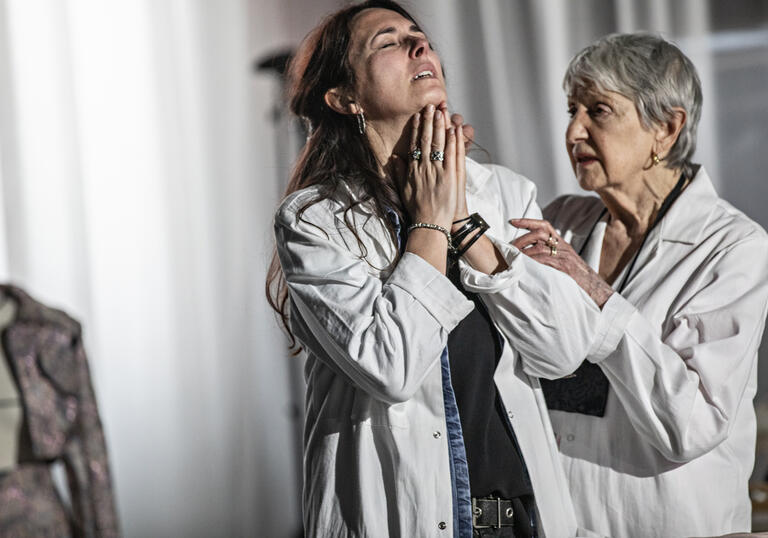Two people in lab coats against a white backdrop where one is comforting the other