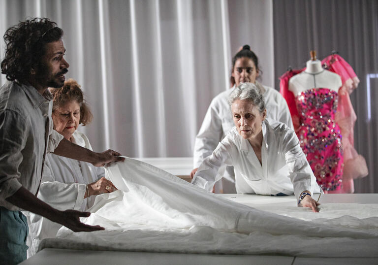 Image of a group of people in lab coats stood around a desk with a white cloth on top of it, with a mannequin wering a pink dress behind them 