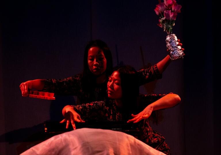 Two women pose theatrically; one holds flowers above, the other draped in white fabric.