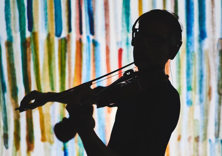 A silhouette of a violinist in front of a multi colour background