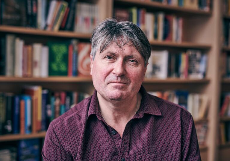 Simon Armitage poses for the camera in front of a bookcase filled with books.