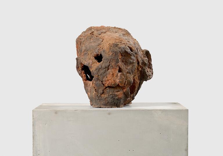 A terracotta sculpture that resembles a human head, sitting on a concrete plinth