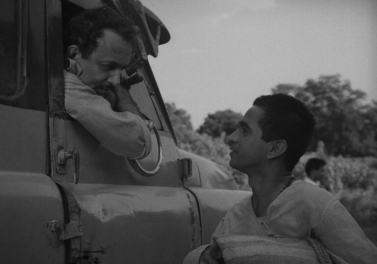 A black and white still from Badnam Basti, showing a man leaning out of a truck window speaking to another man.