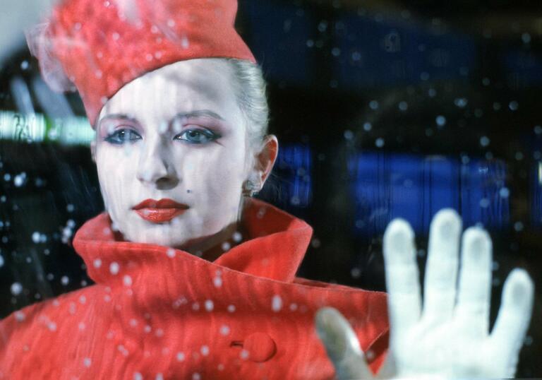 A woman in a red coat and red hat stands in front of a window with her hand placed up on the glass.
