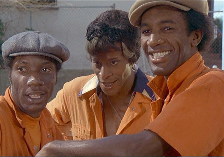 Three people in orange overalls chat together and stare into the camera