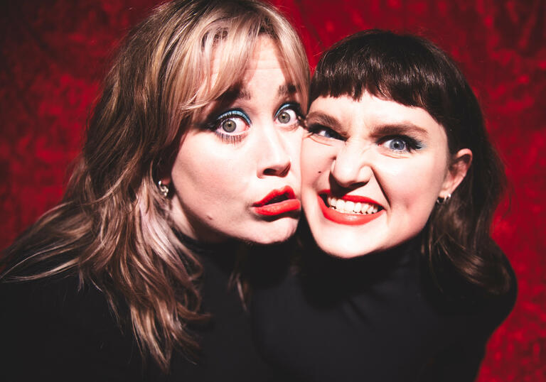 Two people wearing black against a red background, smiling with cheeks pressed together