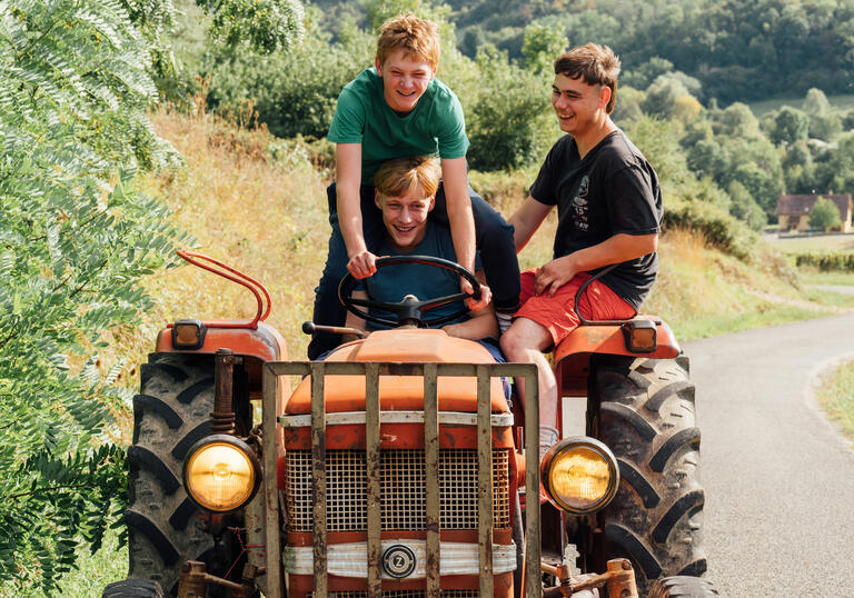 Three teenagers ride on top of a red tractor, grinning