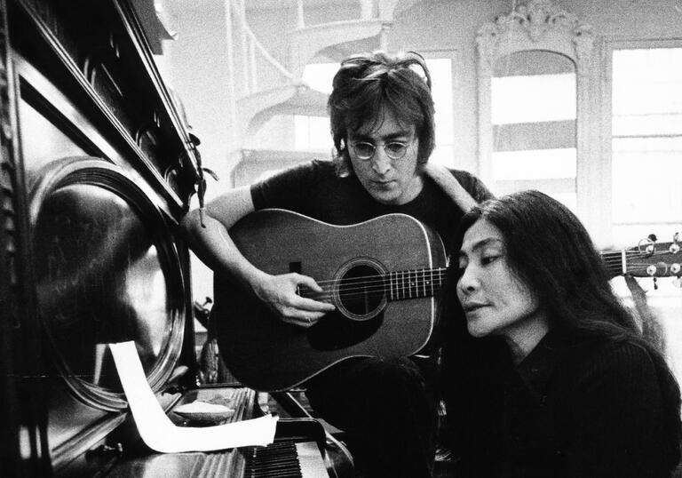 John Lennon and Yoko Ono sit at a piano, reading music in their New York apartment