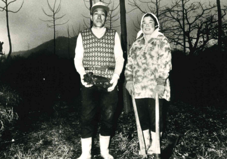 Two people smile at the camera, waring outdoor clothing in a black and white photograph
