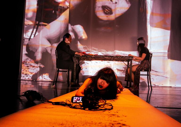 Woman on orange-lit stage with camera; large projected face creates surreal, haunting atmosphere.