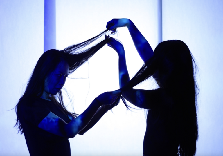 Two silhouetted figures interact with each other's hair against a bright, blue-toned background.