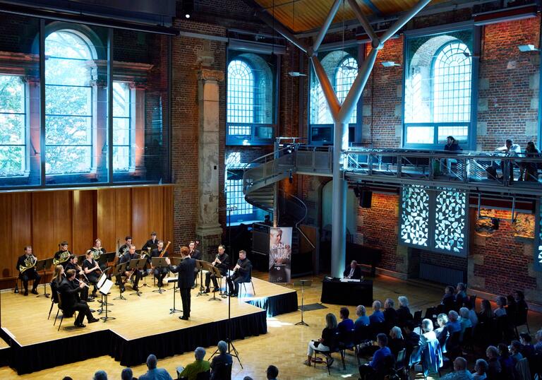 A view of the Jerwood Hall in LSO St LUke's from the balcony, with a chamber group of LSO musicians sat performing on the stage, an audience watching, and sun coming through the venue windows