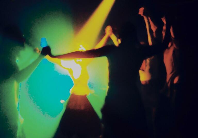 Figures are dancing in a dark room lit by yellow, green and blue lights.