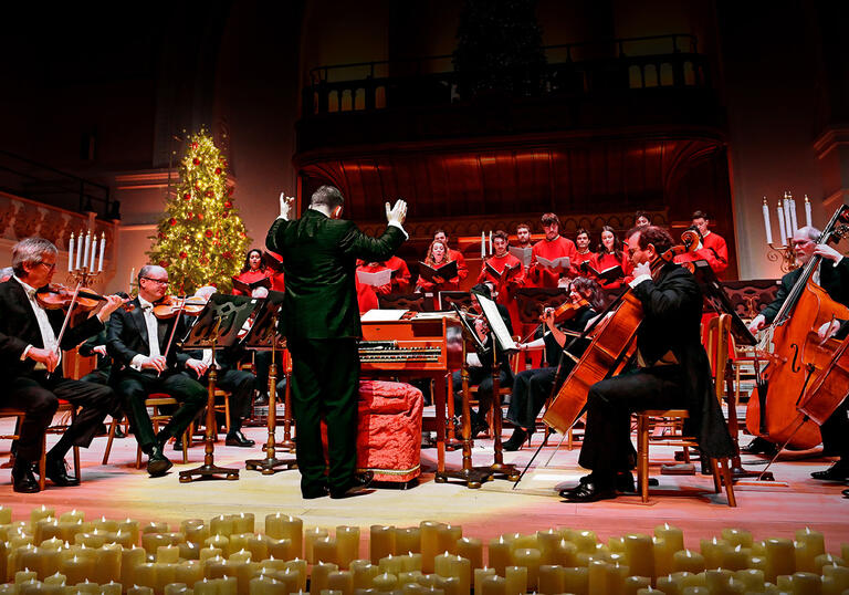 conductor conducting a choir and orchestra behind a row of candles