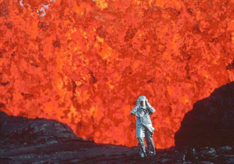 A person in a protective suit standing in front of an erupting volcano.