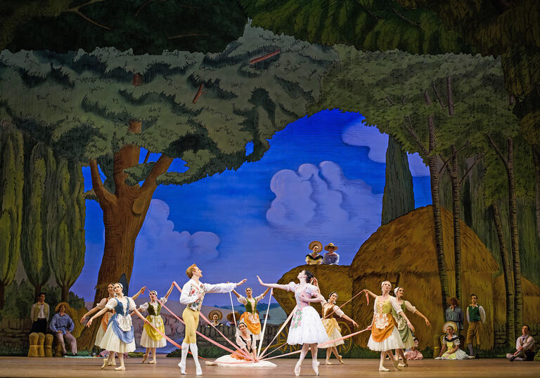 Ballet dancers on stage with ribbons, set against a countryside painted background