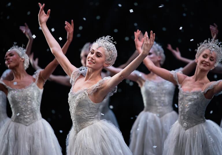 Ballet dancers dressed as snowflakes dance with their arms raised, snow falling around them