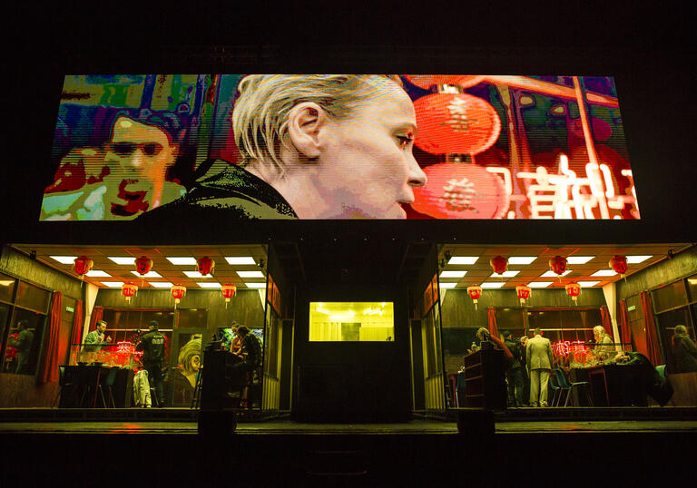 People move around in a chinese restaurant as a close-up image of one person in the venue is projected on a large screen above the set.