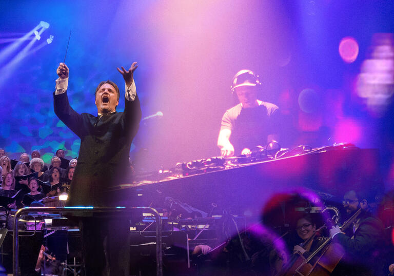 Conductor Fernando Furones leads an orchestra in the foreground with an image of a choir on the left hand side and an image of Symphonic standing behind a DJ deck on the right hand side