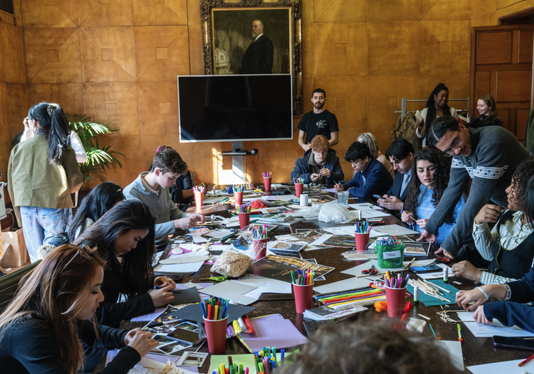 A room of young people sit at a large table with lots of paper, pens and pencils, working together to come up with design ideas