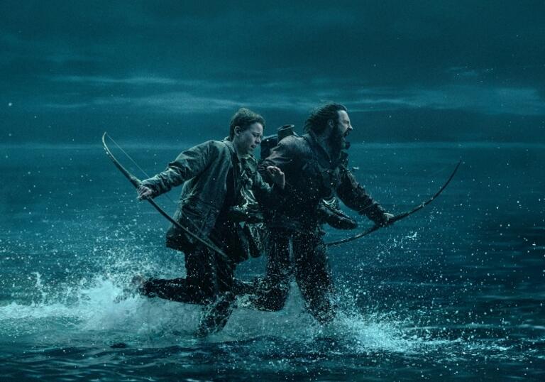 Two people run through water in the dark, holding a bow and arrow