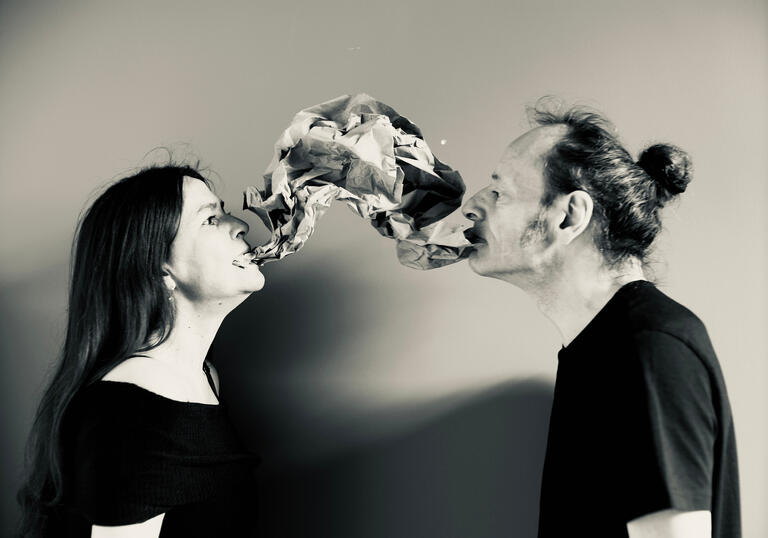 Two people stand opposite each other, a material shape - part balloon, part speech bubble - hangs between them.