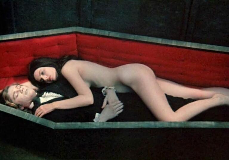 A naked woman lies on a man in a suit in a coffin.