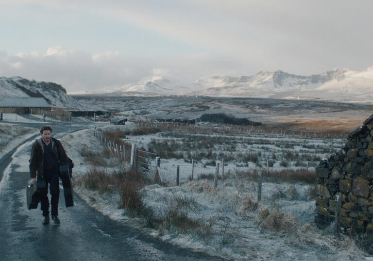 A man walks down a snowy path, holding lots of luggage