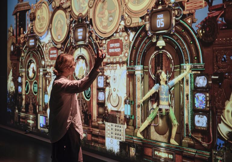 A woman stands in front of a large wall projection, mimicking the dance move shown by the character on the screen