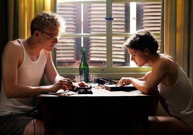 A blonde man, and brunette woman sit in white vests, smoking and talking across a table