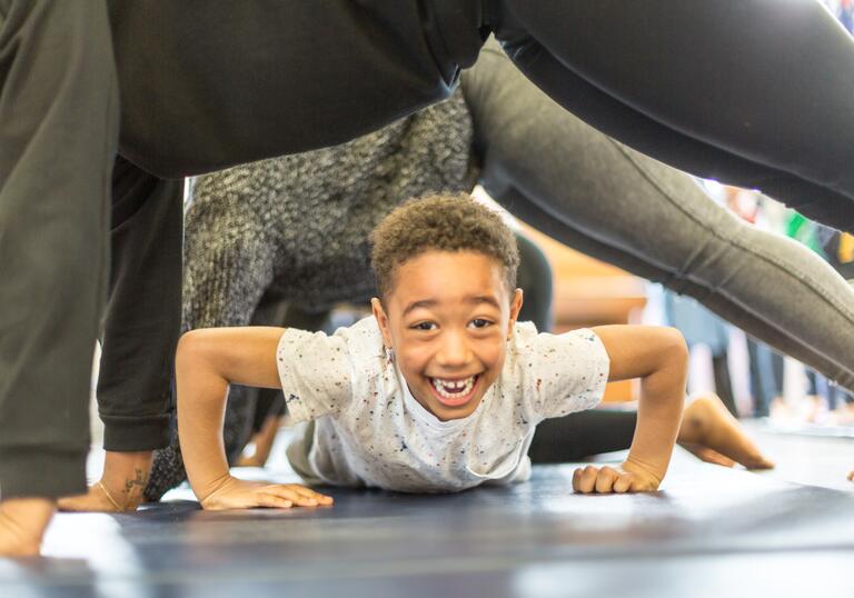 A child smiling and crawling on the ground underneath people who are planking