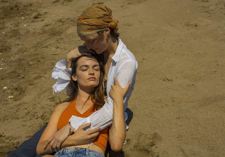 Two women sit on the beach, in each others arms